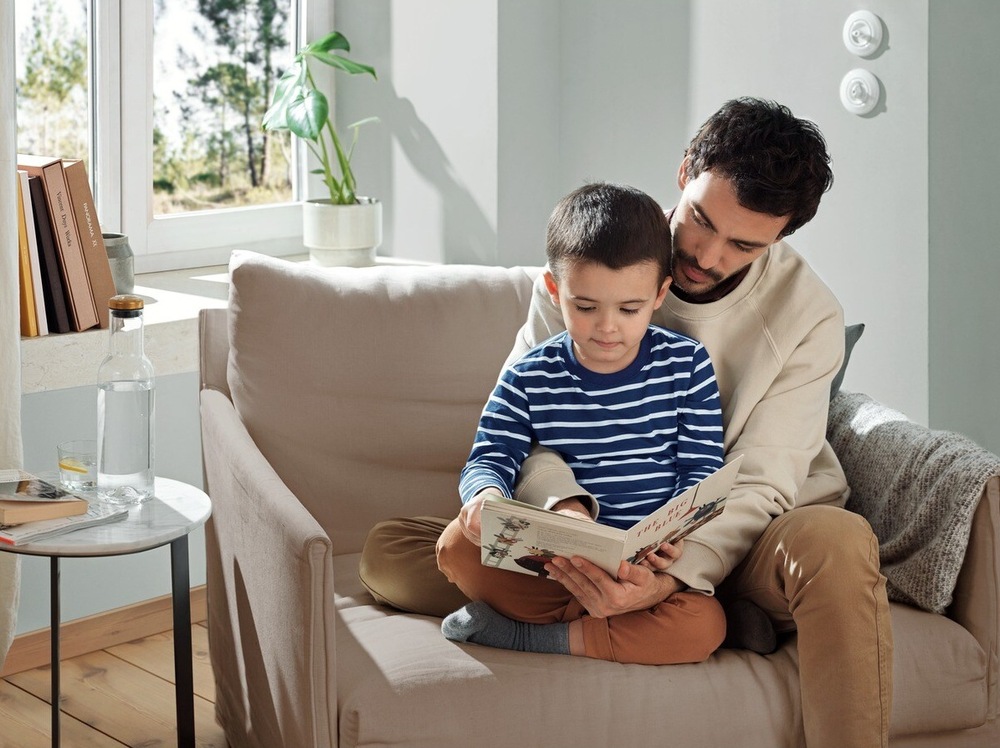 Pračka Electrolux PerfectCare 700 EW7TN23372C bílá Muž sedí s chlapcem v křesle a čtou spolu knihu. V pozadí je okno do zahrady a před ním na parapetu knihy a pokojová květina. Vlevo vedle křesle je stolek s lahví a sklenicí vody.