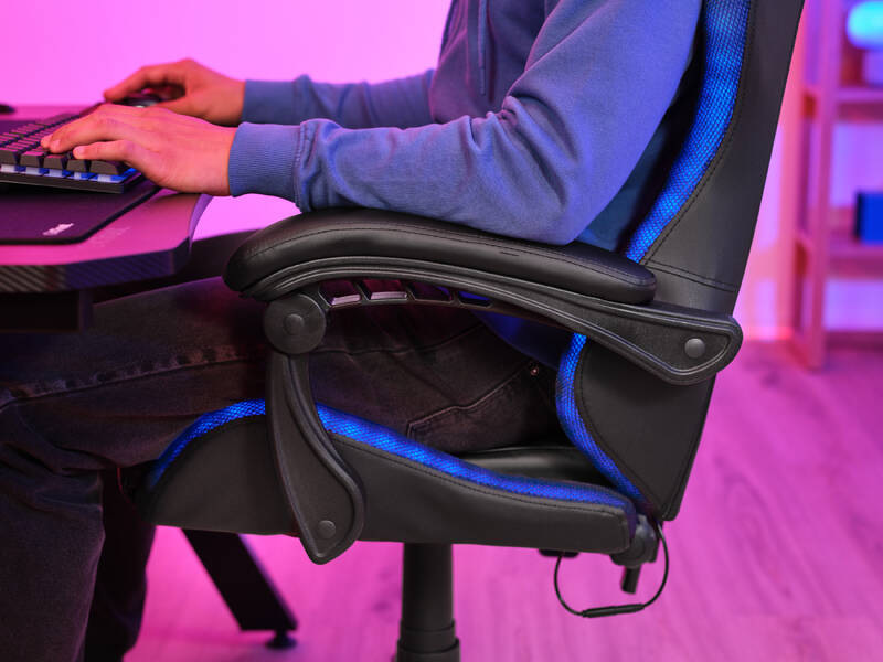 V herní židli  Trust GXT 704 RONIQ RGB sedí osoba v modré mikině a hraje hru, židle s RGB osvětlením po obvodu svítí modře. 