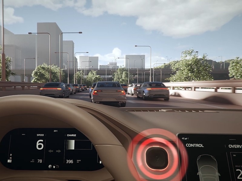 Pohled z interiéru automobilu na kolonu pomalu jedoucích vozidel. Cestovní asistent TomTom Tom na palubní desce svítí červeným LED kroužkem a upozorňuje řidiče na zpomalený provoz před vozem.