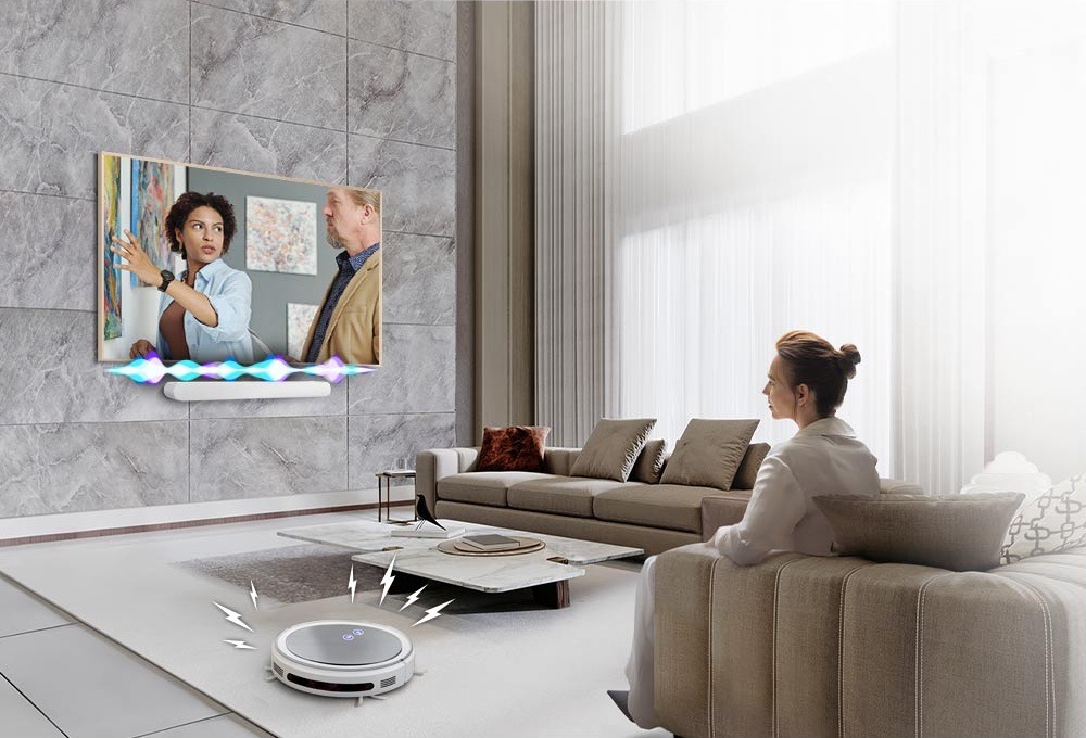Soundbar Samsung HW-S61D bílý Soundbar Samsung je připevněn na zdi pod televizí Samsung, na jejíž obrazovce spolu hovoří muž a žena, nad soundbarem je nákres proměnlivé intenzity zvuku, v popředí je žena sledující televizi na pohovce a vlevo od pohovky je robotický vysavač