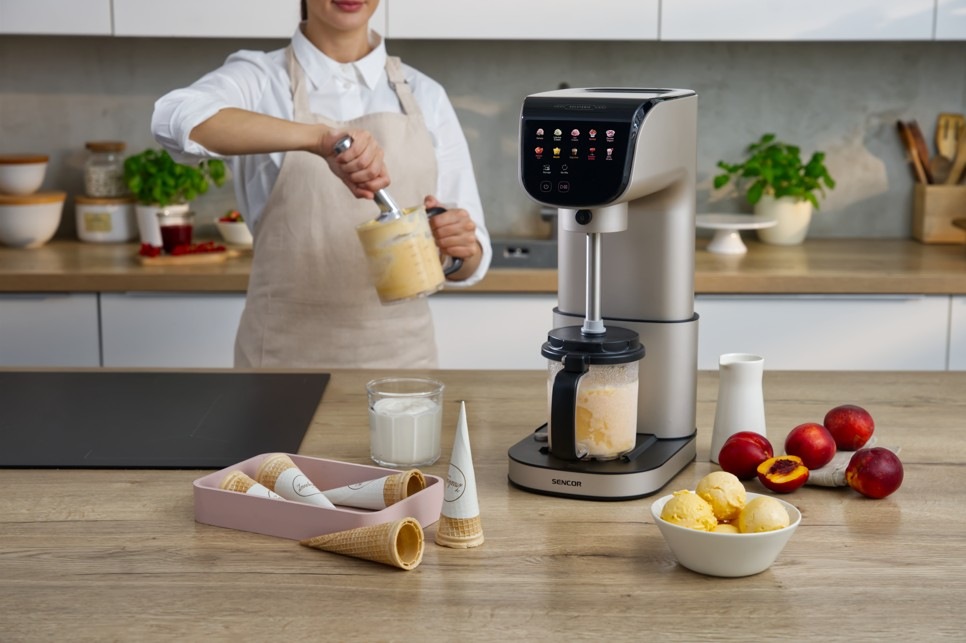 Zmrzlinovač Sencor SIC 5050WS GELATERIA champagne Žena připravuje krémovou zmrzlinu pomocí zmrzlinovače Sencor SIC 5050WS GELATERIA v moderní kuchyni. V nádobě se míchá světlá směs, zatímco na pracovní desce jsou připravené kornouty, mléko a čerstvé broskve. Vedle stojí miska s hotovými kopečky zmrzliny.