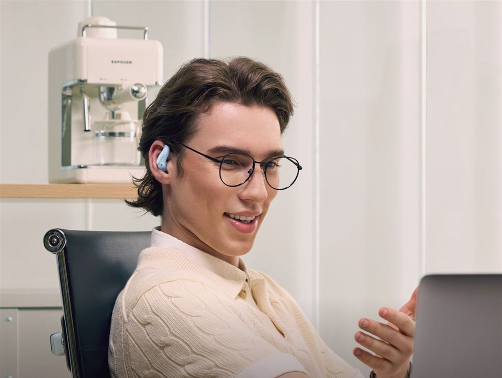 Sluchátka Shokz OpenFit Air 2, Bluetooth modrá Muž v brýlích sedí v kanceláři u pracovního stolu, má nasazené sluchátka OpenFit Air 2, v ruce drží telefon a s někým hovoří.