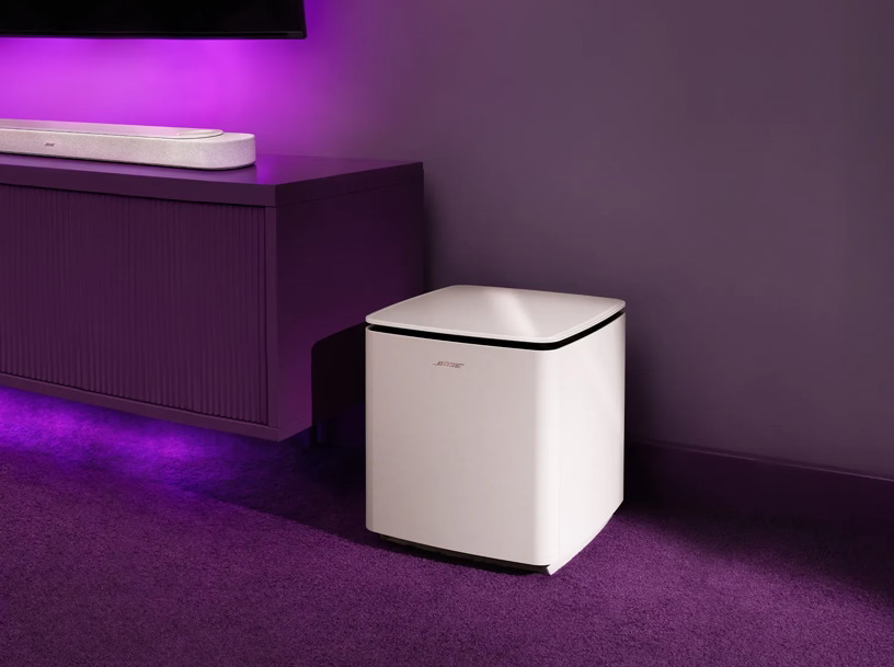 Subwoofer Bose Bass Module umístěný na podlaze u TV stolku s fialovým podsvícením.
