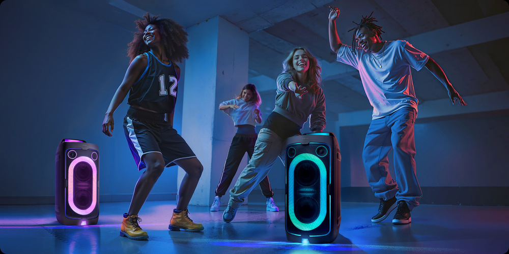 Dva reproduktory Samsung MX-ST40F stojí na zemi, jeden svítí růžově, druhý zeleně, kolem nich tančí čtyři lidé