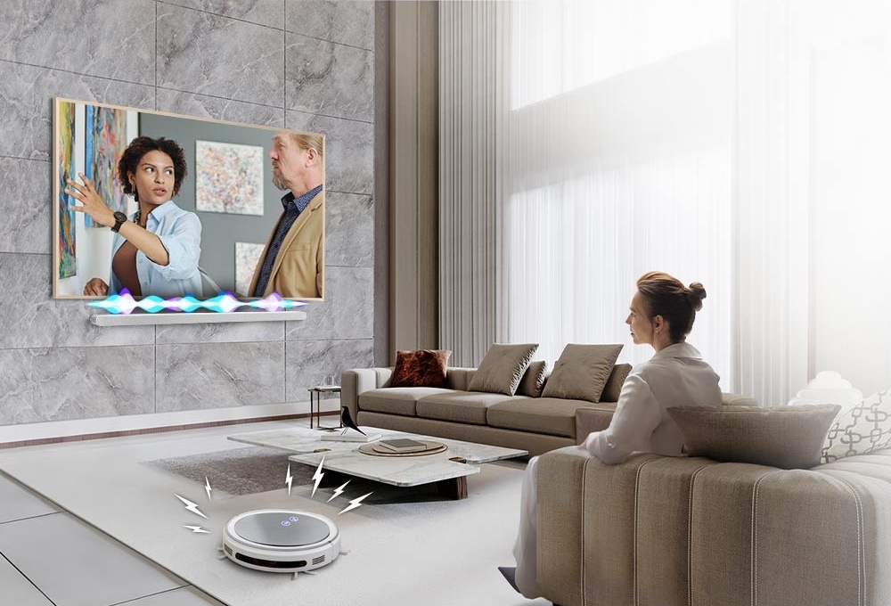 Samsung HW-S801D je připevněn na zdi obývacího pokoje pod televizí Samsung, nad soundbarem vlnky znázorňují hlasitost zvuku, v popředí sedí žena sledující televizi a vedle ní je robotický vysavač s čárami znázorňujícími hluk