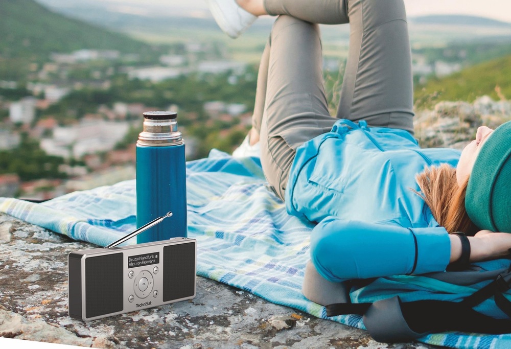 Radiopřijímač s DAB+ TechniSat DIGITRADIO 1 S černý/stříbrný Radiopřijímač TechniSat DIGITRADIO 1 S s vysunutou anténou stojí na skále vedle termosky. V pozadí odpočívá turistka na dece s výhledem do krajiny, což znázorňuje použití rádia na cestách.