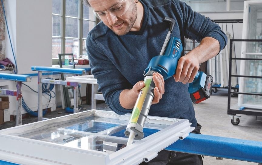 Lis Bosch Professional GCG 18V-310 (bez baterie) Muž v dílně používá vytlačovací pistoli Bosch na silikon a utěsňuje skleněnou výplň na okně.