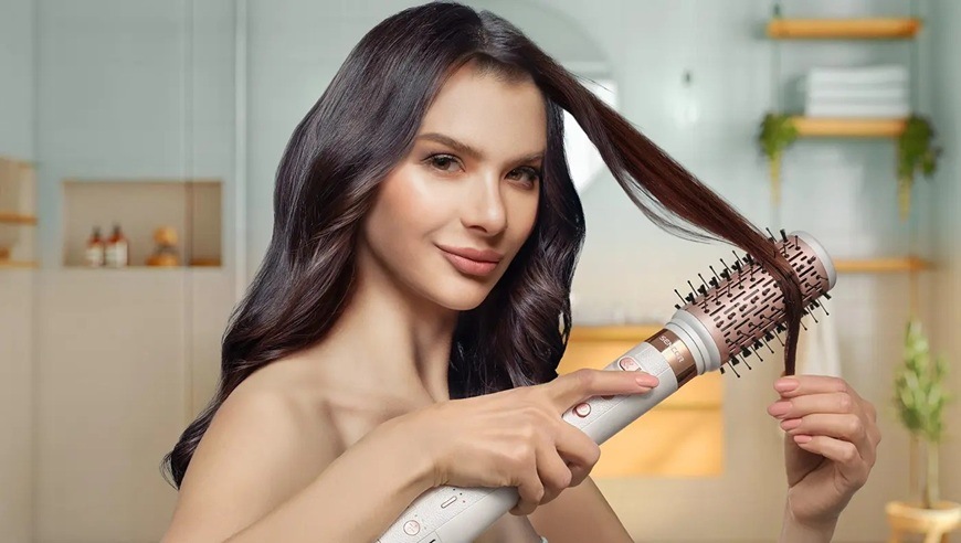 „Žena s dlouhými tmavými vlasy používá multistyler Sencor SHS 9200RS s kulatým kartáčovým nástavcem, kolem kterého je omotaný pramen vlasů. Jednou rukou drží přístroj s ovládacími tlačítky, druhou upravuje vlasy.