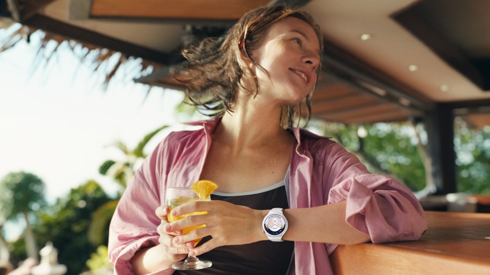 Chytré hodinky Huawei Watch 6 42 mm zlaté Usmívající se žena s mokrými vlasy odpočívá u baru s nápojem v ruce; na zápěstí má chytré hodinky WATCH 6 s elegantním analogovým ciferníkem