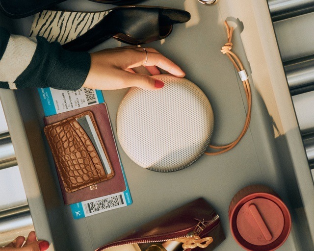 Přenosný reproduktor Bang & Olufsen Beosound A1 (3rd Gen) červený Přenosný reproduktor Bang & Olufsen A1 (3rd Gen), červený leží v přihrádce s věcmi ke kontrole na letišti, k reproduktoru se natahuje ženská ruka