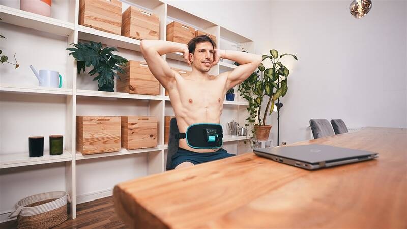Elektrostimulátor Beurer Corefit II černý Muž sedí v pokoji u stolu a používá břišní pás Beurer Corefit II