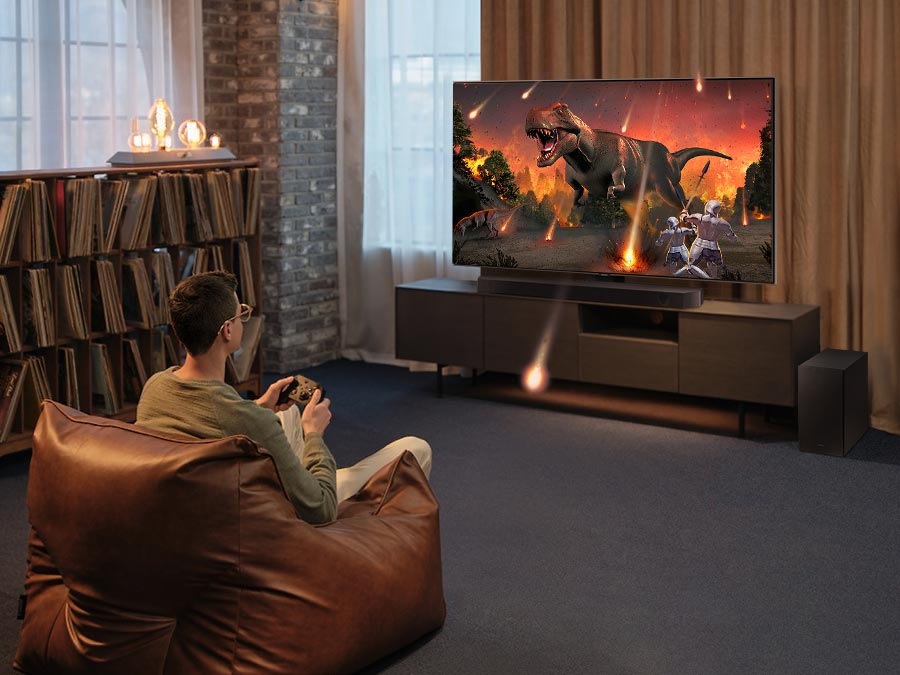 Muž sedí na sedacím vaku na zemi před televizí a soundbarem Samsung HW-Q600C na televizním stolku a hraje videohru, jiskry z obrazu videohry přecházejí mimo TV