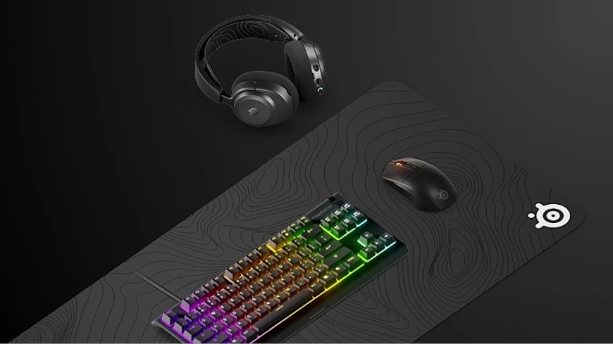 Podložka pod klávesnici a myš SteelSeries QcK XXL Topographic černá Podložka QcK XXL Topographic pod klávesnicí a myší, vedle ní headset na stole