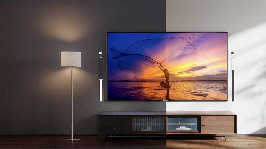 Na obrázku je televize Hisense umístěná v obývacím pokoji, zobrazující surfaře při západu slunce. Scéna je rozdělena na dvě poloviny – jedna znázorňuje temnější večerní osvětlení a druhá jasné denní světlo, čímž ilustruje funkci adaptivního světelného senzoru, který automaticky upravuje jas obrazovky podle okolních podmínek.