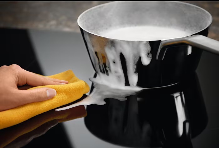 Indukční varná deska Electrolux Inspiration EIV634 černá Skleněný povrch indukční desky s rendlíkem s přetékajícím mlékem. Ruka utírá rozlité mléko z desky hadříkem.