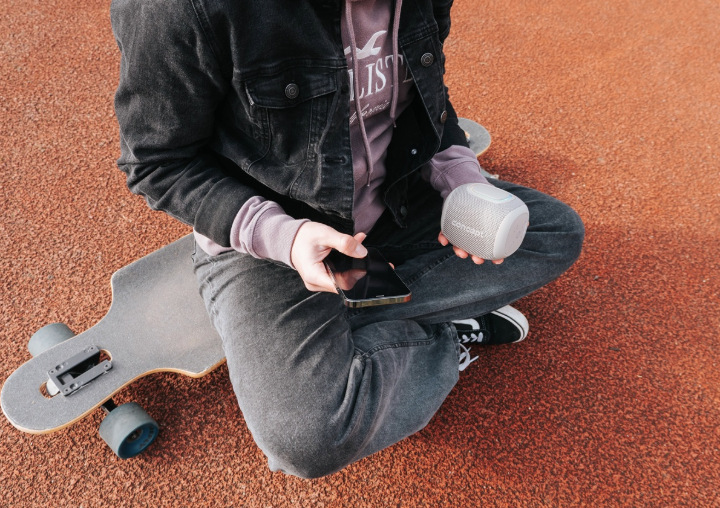 Uživatel drží reproduktor Concept SoundJoy BR5011 a telefon při posezení vedle longboardu