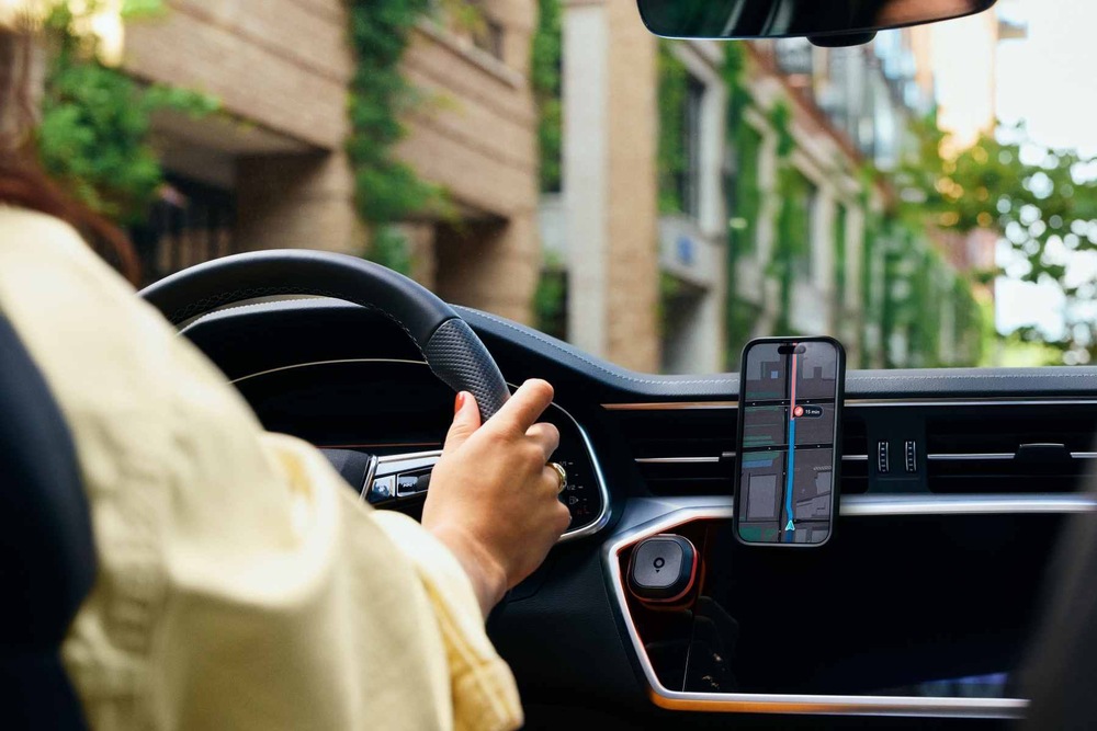 Řidička sedí za volantem automobilu, zatímco telefon upevněný na palubní desce zobrazuje navigační trasu v aplikaci TomTom. Pod telefonem je na magnetickém držáku umístěný cestovní asistent TomTom Tom.