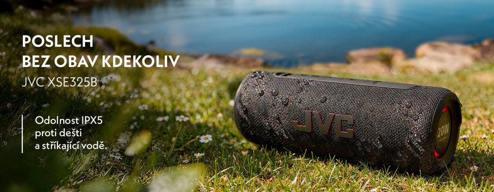 Černý voděodolný reproduktor JVC XSE325B s kapkami vody na povrchu v trávě u jezera. Text: „Poslech bez obav kdekoliv – Odolnost IPX5 proti dešti a stříkající vodě“.