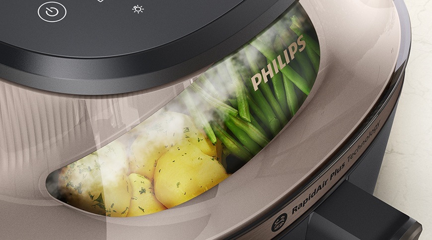 Detail horkovzdušné fritézy Philips NA547/07 s průhledným oknem, za kterým je vidět připravovaná zelenina, včetně brambor a zelených fazolek. Na povrchu spotřebiče je viditelné logo Philips a označení technologie RapidAir Plus.