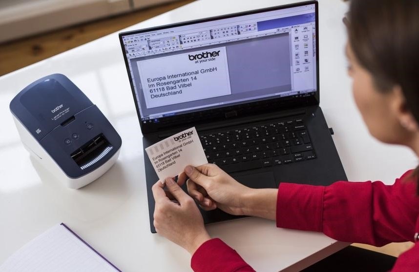 Tiskárna štítků Brother QL-600 bílá/modrá žena sedí u počítače, na kterém připravila štítek dle požadavků, vedle je na stole položená také tiskárna štítků Brother QL-600 s vytištěou etiketou.