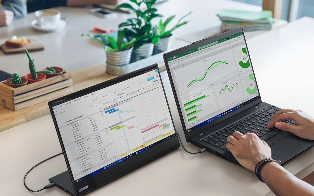 Notebook Lenovo ThinkPad L14 Gen 6 černý Na stole jsou dva notebooky, na kterých jsou zobrazeny grafy a plány, obklopené malými rostlinami v květináčích, a osoba, která píše na klávesnici, vytváří soustředěnou pracovní atmosféru.