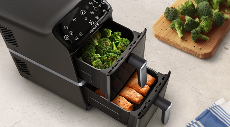 Fritéza horkovzdušná Philips Airfryer Dual Basket 4000 series 10 l, NA461/00 černá Horkovzdušná fritéza Philips NA461/00 se dvěma vysunutými koši, v horním koši je brokolice a ve spodním filety lososa, vedle na kuchyňské lince je dřevěné prkénko s další zeleninou.