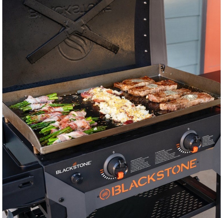 Blackstone 28" Omnivore na verandě u modré fasády s připojenou propanovou lahví.