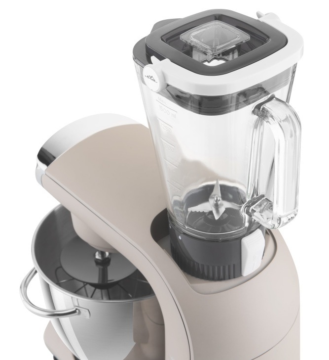 Detail skleněného mixéru o objemu 1,5 l nasazeného na horním vývodu kuchyňského robota ETA Gratus EVO Smart II. Pohled na nerezové nože mixéru a odměrnou stupnici, v pozadí nerezová mísa