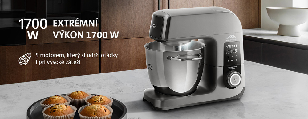 Titanový kuchyňský robot ETA Gratus Kuliner II Vario s nerezovou mísou v moderní kuchyni, vedle talíře s muffiny. Text v obrázku zdůrazňuje extrémní výkon 1700 W a motor se stabilizací otáček při vysoké zátěži