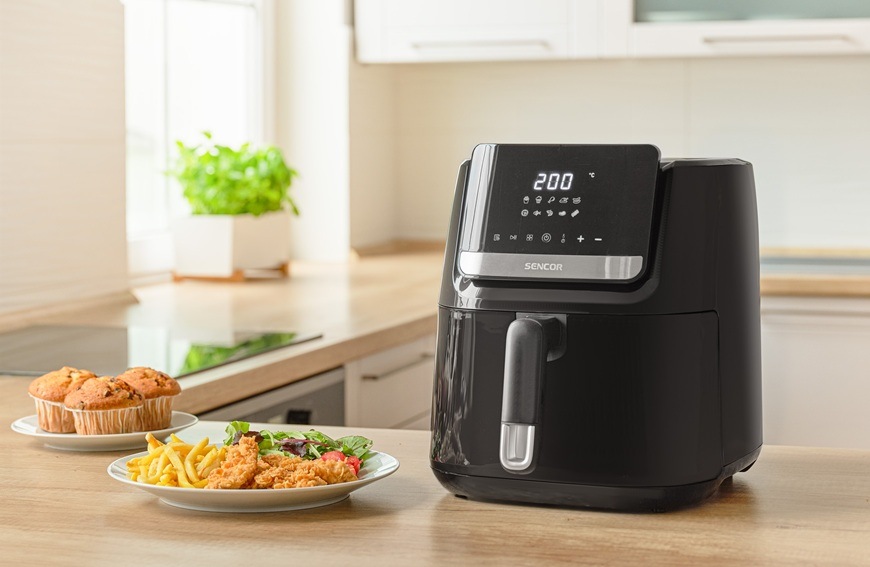 Fritéza horkovzdušná Sencor SFR 6550BK-EUE3 černá Horkovzdušná fritéza Sencor SFR 6550BK-EUE3 je položená na světlé kuchyňské lince. Vedle ní jsou položené stripsy s hranolkami, zeleninou a muffiny.