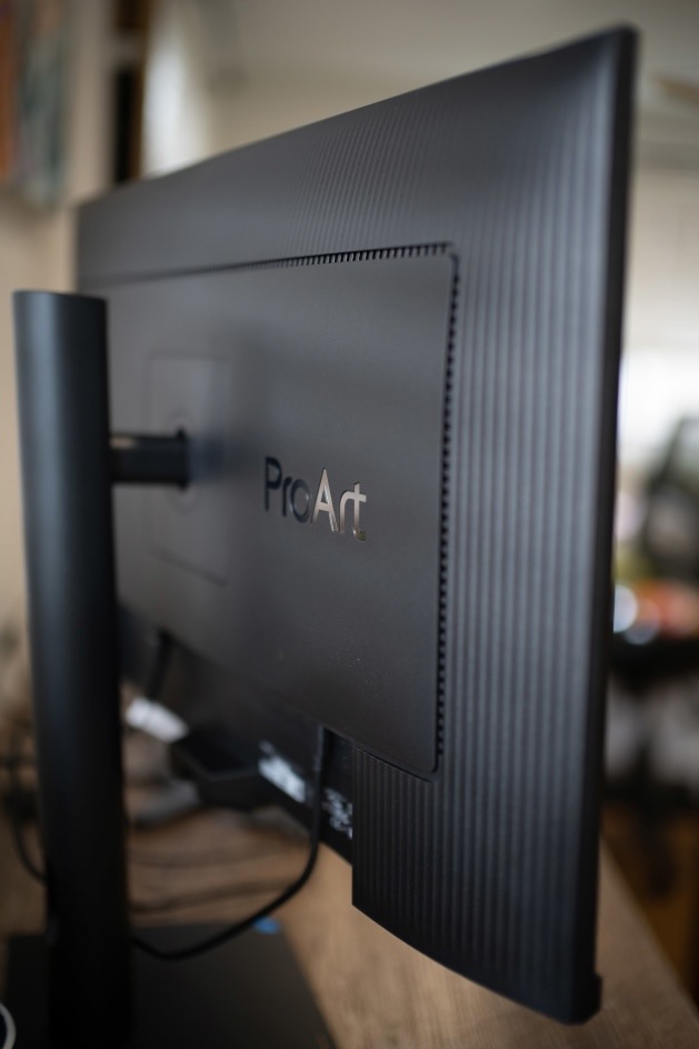 Monitor Asus ProArt PA27UCGE černý Na obrázku je zadní strana monitoru Asus ProArt PA27UCGE 27" zachycená z bočního detailu. Monitor má černý zadní kryt s jemným svislým žebrováním a uprostřed plastový panel s logem ProArt. Vlevo je vidět svislý stojan a rameno, které drží displej. Ve spodní části zadní strany jsou připojené kabely. Snímek zdůrazňuje zadní provedení monitoru, jeho uchycení ke stojanu a profesionální matný design.