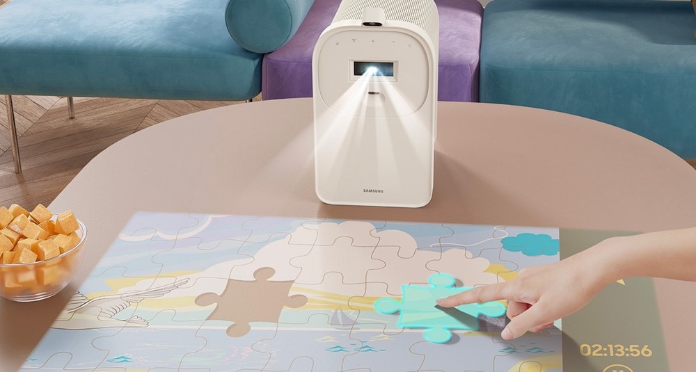 Laser TV Samsung SP-LPF5DSA Projektor The Premiere 5 ve stojánku stojí na stole a promítá na něj puzzle, lidská ruka posouvá dílek puzzle