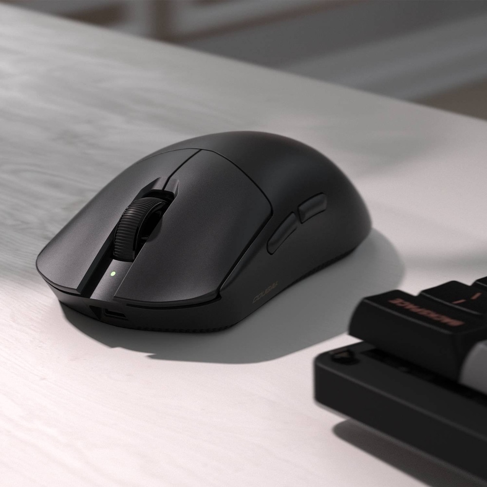 Na obrázku je černá bezdrátová herní myš Cougar Revenger PRO 4K položená na světlém stole vedle klávesnice. Myš má matný povrch, výrazně klenutý ergonomický tvar, vroubkované rolovací kolečko a malou zelenou stavovou kontrolku. Na levém boku jsou vidět dvě boční tlačítka pro palec. V přední části je otvor pro připojení kabelu k nabíjení nebo kabelovému použití.