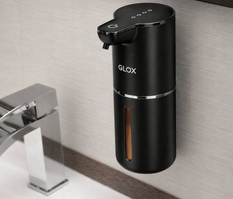 Automatický bezdotykový dávkovač mýdla GLOX GM1B v černém provedení, který je prostřednictvím oboustranné lepicí pásky přilepen na zeď vedle umyvadla.