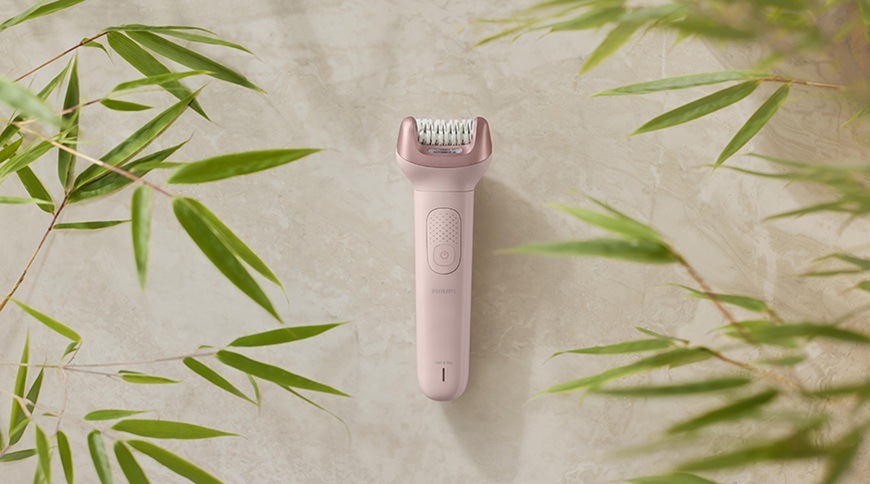 Epilátor Philips BRE718/00 v růžové barvě položený na světlém povrchu, kolem něj jsou dekorativní zelené listy a zařízení je zobrazeno zepředu.