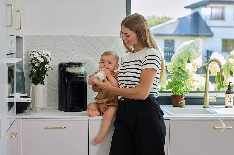Maminka posadila své děťátko na kuchyňskou linku vedle , příčemž v každém je umístěna velikostně jiná lahvička ohřívače a krmí ho právě připravenou lahvičkou.