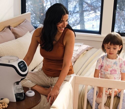 Maminka má vedle postele postavený ohřívač mléka Baby Brezza Formula Pro Mini a chystá pro miminko v postýlce kojeneckou lahvičku.