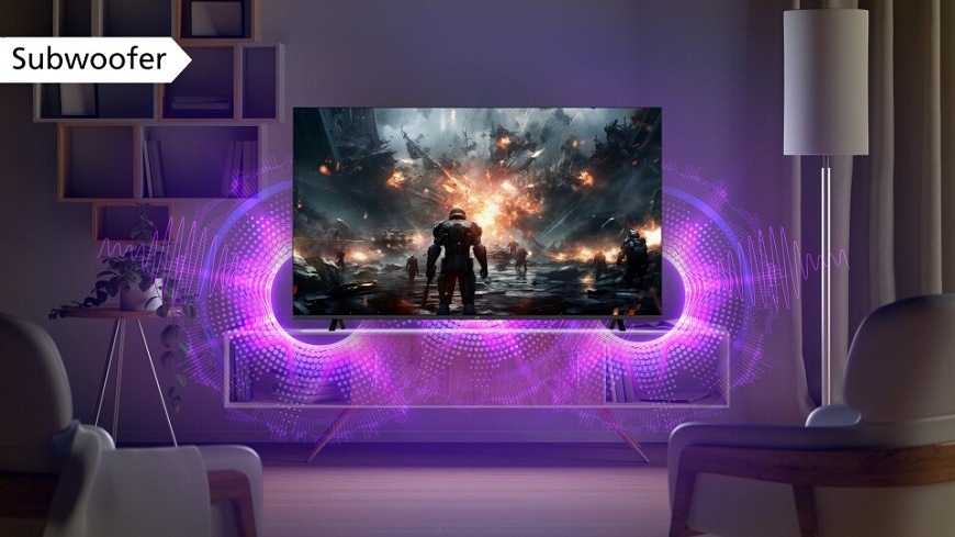 Televize JVC umístěná v moderním obývacím pokoji, na jejíž obrazovce je zobrazená scéna z akční videohry s futuristickým vojákem. Z dolní části televizoru jsou fialovými vlnami zvýrazněny zvukové efekty, které naznačují silný basový výkon vestavěného subwooferu. V levém horním rohu je popisek "Subwoofer"