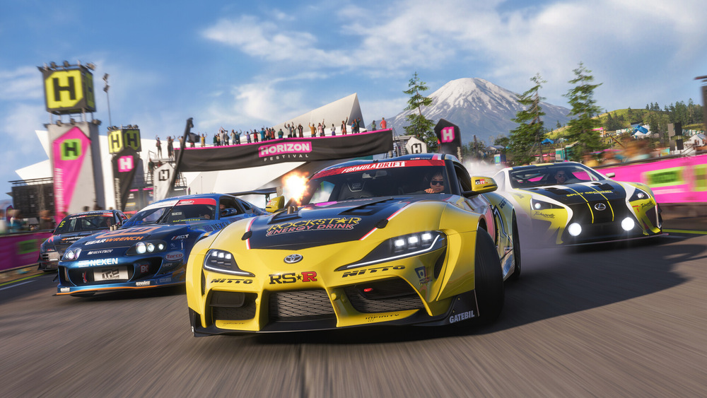 Závodní hra Forza Horizon 6 se záběrem na horské prostředí se třemi sportovními vozy v akci. Živý dav a transparenty vytvářejí vzrušující a dynamickou atmosféru.