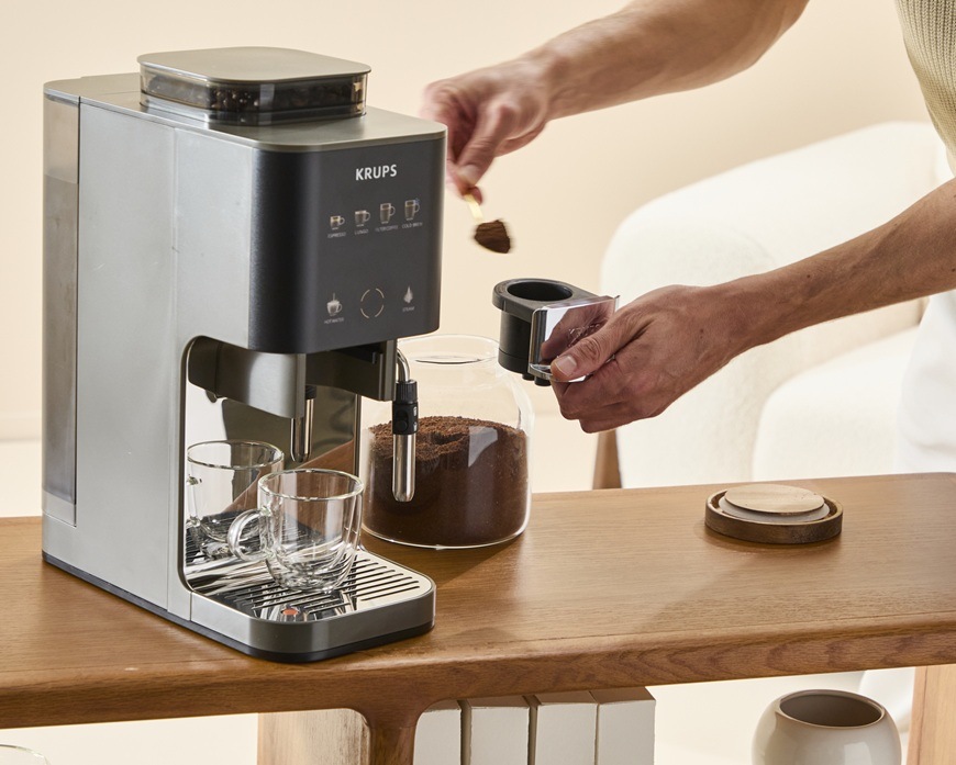 Žena má v ruce lžíci a na ni má mletou kávu. Tu dává do zásobníku v automatickém kávovaru Krups SA403BE0 Coffee Crush Experience.
