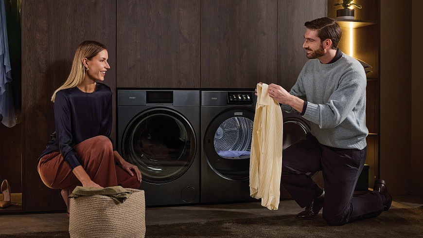 muž držící oblečení a žena klečí u otevřené osvětlené sušičky Haier HD90-BQL8GNU1-S a pračky haier v prádelně