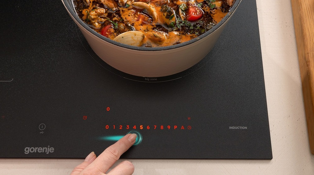 ruka ovládá varnou desku Gorenje GI6433SRWF. Na desce stoji hrnec s hustou polevkou.