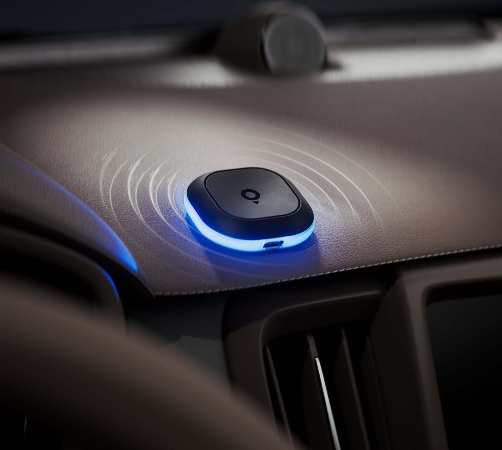 Cestovní asistent TomTom Tom položený na palubní desce automobilu. Modře svítící obvodový LED kroužek a znázorněné zvukové vlny signalizují upozornění na rychlostní kameru.