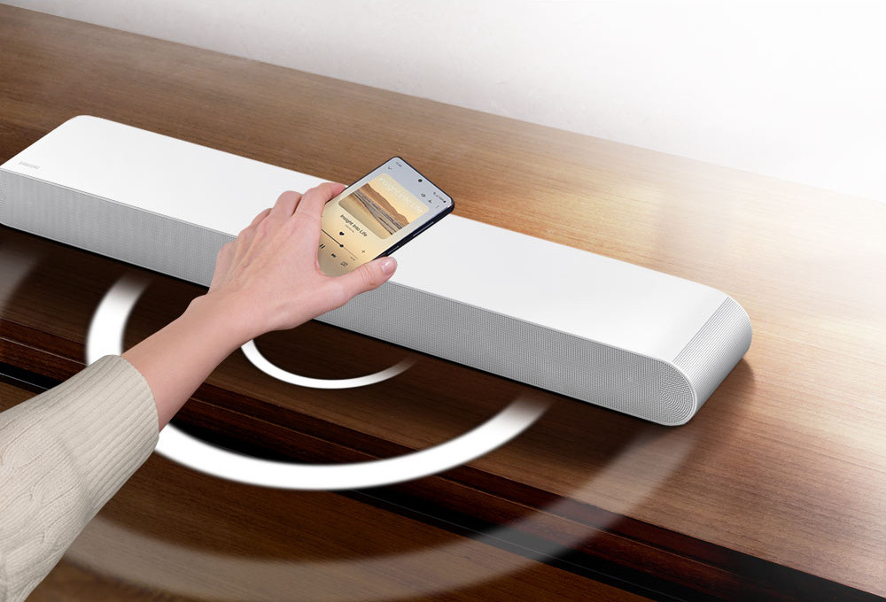 Soundbar Samsung HW-S61D bílý Ruka drží mobilní telefon přehrávající hudbu nad bílým soundbarem Samsung HW-S61D, z nějž vycházející bílá kolečka znázorňující zvuk