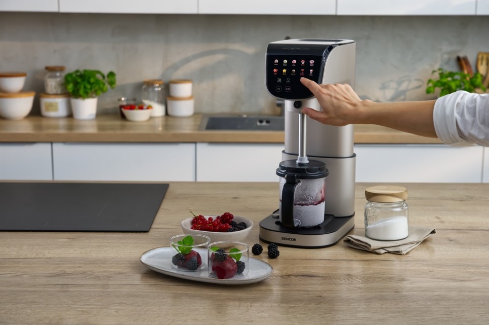 Zmrzlinovač Sencor SIC 5050WS GELATERIA champagne Zmrzlinovač Sencor SIC 5050WS GELATERIA v moderní kuchyni během přípravy ovocného sorbetu, kdy uživatel volí program na dotykovém displeji. V nádobě se míchá směs z lesního ovoce a na pracovní desce jsou připravené čerstvé suroviny i hotové dezerty ve skleničkách. Celá scéna působí čistě a elegantně.
