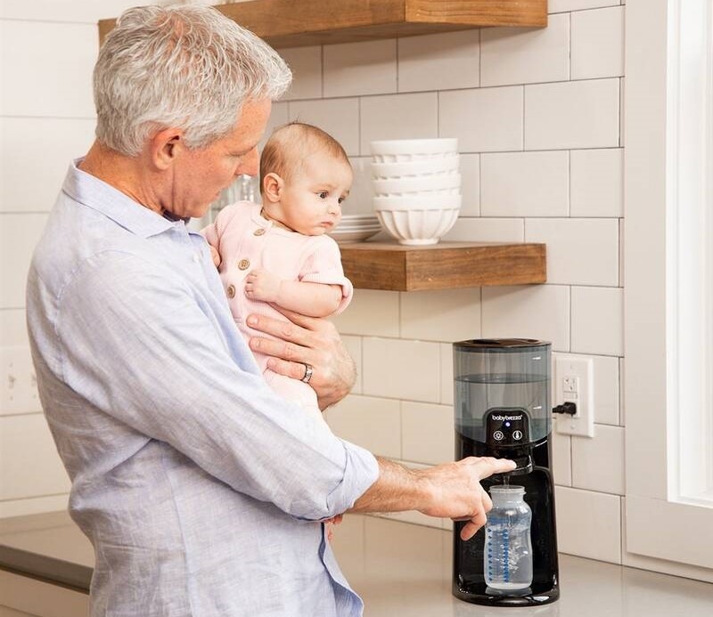 Pán drží v náručí miminko a přitom obsluhuje ohřívač vody Baby Brezza Instant Warmer Black.