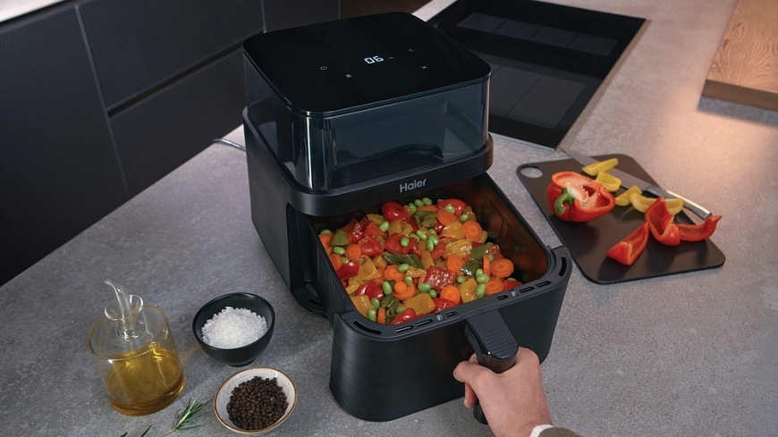 Fritéza horkovzdušná Haier I-Master Series 3 HAF3AFW 011 černá Horkovzdušná fritéza Haier HAF3AFW 011 na kuchyňské lince, vedle ní se nachází ingredience, uvnitř fritovacího koše je směs zeleniny