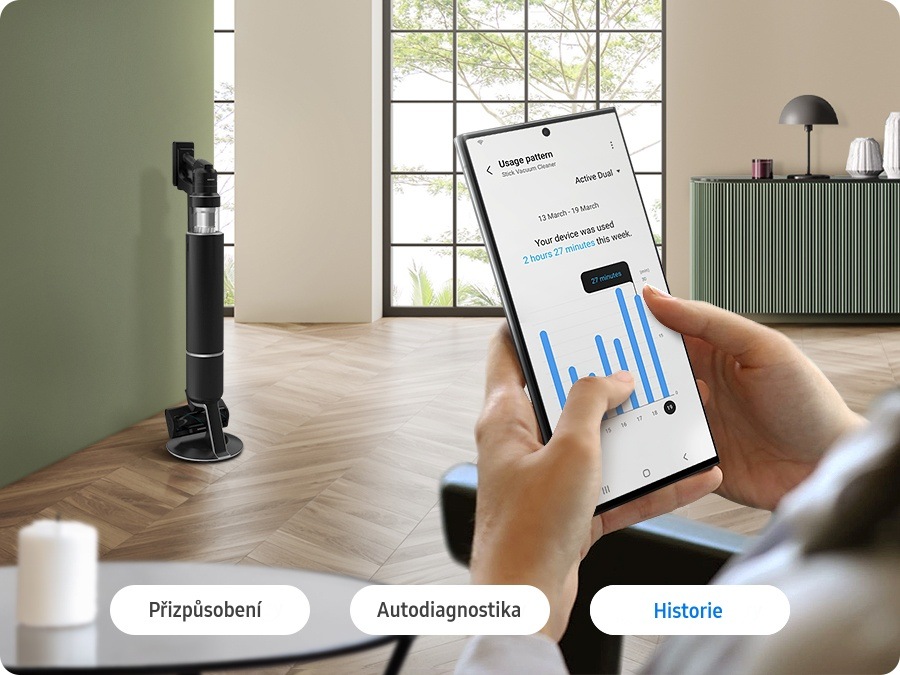 Ruce drží mobilní telefon, na jehož displeji je aplikace SmartThings, na pozadí je tyčový vysavač Samsung BESPOKE JET AI VS28C9784QK/GE v obývacím pokoji