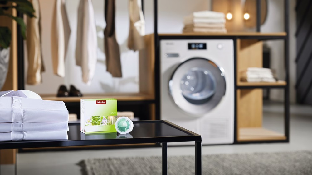 Vůně do sušičky Miele Nature, 12,5 ml Miele FA N 151 L flakón s vůní Nature položený na stole vedle složených košil, sušička Miele v pozadí