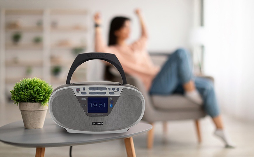 Radiomagnetofon s DAB+/CD AIWA BBTU-500DAB stříbrný Radiopřijímač DAB+/CD Aiwa BBTU-500DAB/SL je položený na konferenčním stole. Žena v pozadí sedí v křesle a poslouchá svou oblíbenou hudbu.